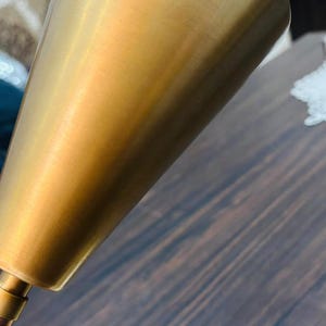 May include: A brass-coloured metal lamp shade with a conical shape and a smooth, brushed finish. The shade is attached to a thin, matching metal arm that connects to a circular base, featuring a clean, metallic aesthetic throughout the design.