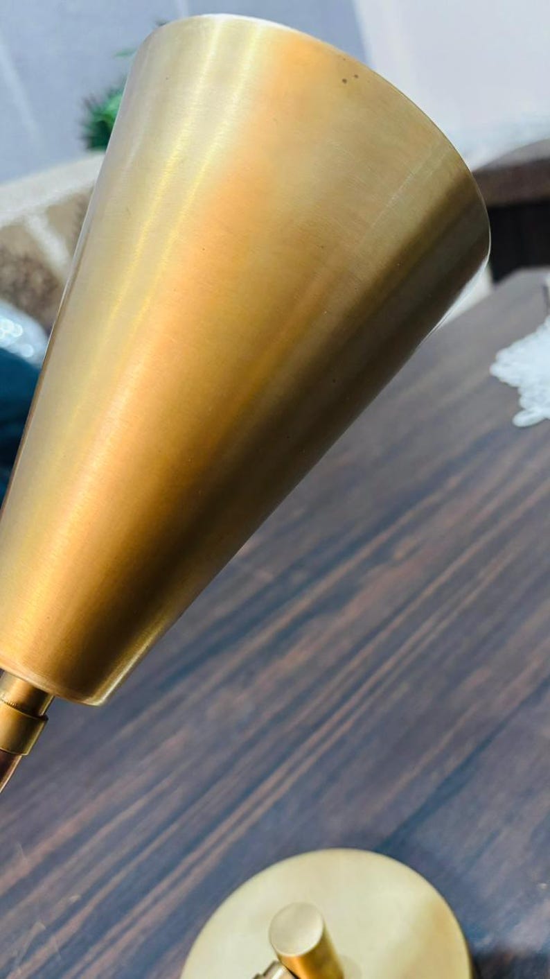May include: A brass-coloured metal lamp shade with a conical shape and a smooth, brushed finish. The shade is attached to a thin, matching metal arm that connects to a circular base, featuring a clean, metallic aesthetic throughout the design.