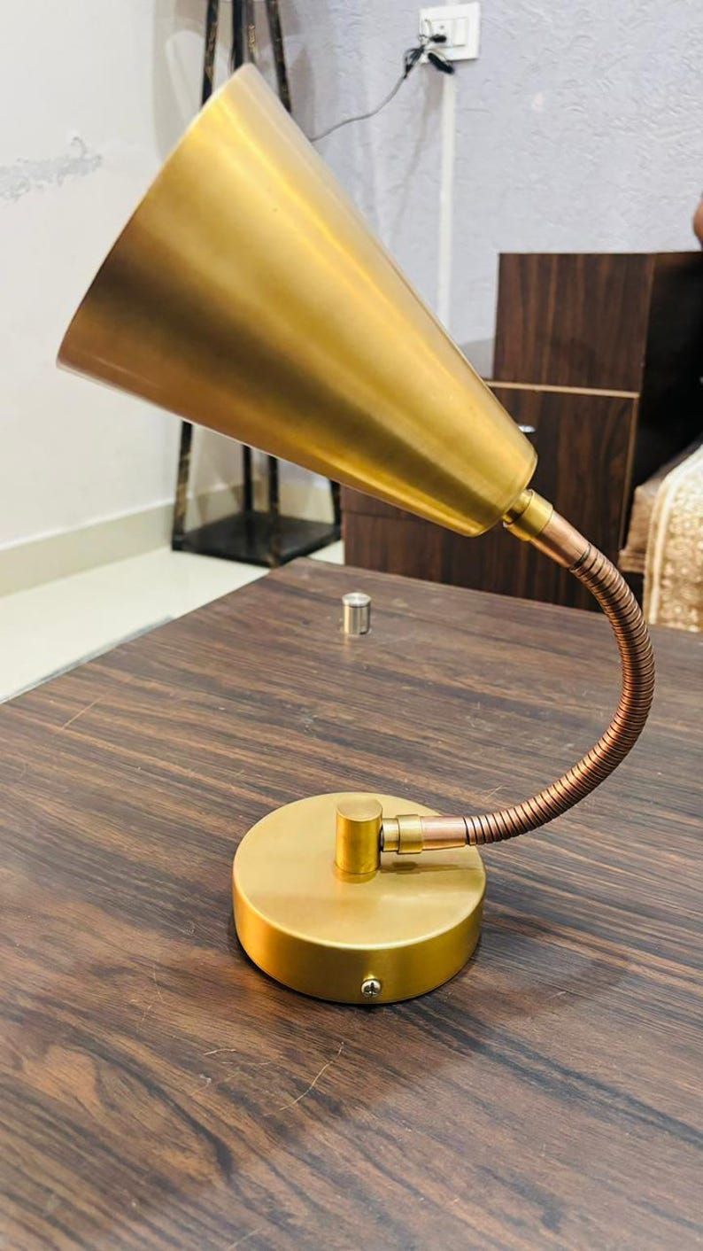 May include: A desk lamp featuring a conical gold-toned metal shade attached to a flexible, ribbed copper-coloured neck. The lamp is supported by a circular gold-toned base with a visible screw on the side, providing a stable foundation for the fixture.