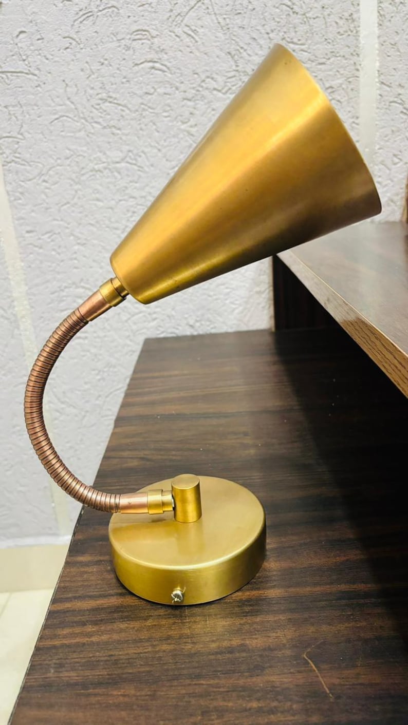 May include: A desk lamp featuring a conical brass shade, a flexible copper-toned gooseneck arm, and a circular brass base. The lamp has a metallic finish and a simple, functional design with a single joint connecting the arm to the base.