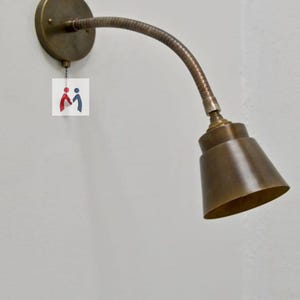 Puede incluir: Una lámpara de pared de tono bronce con un brazo flexible de cuello de cisne. La lámpara tiene una base circular y una pantalla cónica. El diseño está inspirado en la vendimia, adecuado para leer o iluminar tareas. La lámpara está montada en una pared blanca.