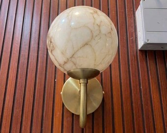 May include: A wall-mounted light fitting featuring a spherical, cream-coloured glass shade with brown marble-like veining. The shade is supported by a brushed brass-toned metal arm and a circular wall plate, creating a clean and functional design.