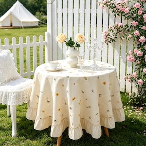 May include: A cream-coloured round tablecloth with a floral pattern, featuring yellow sunflowers and green leaves. The tablecloth has a ruffled edge and is set on a small wooden table. A white chair with lace detailing is on the left.