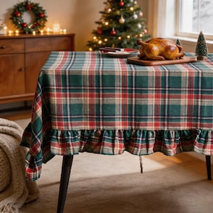 May include: A rectangular tablecloth featuring a classic plaid pattern in shades of forest green, deep red, and cream. The fabric has a gathered ruffle trim along the bottom edge, draping over the sides of a table with dark, slender legs.