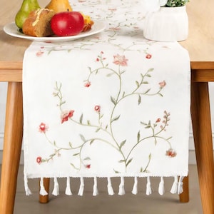 Könnte beinhalten: Weißer Tischläufer mit gestickten rosa Blüten und grünen Stielen. Der Läufer hat eine Fransenborte und wird auf einem Holztisch präsentiert. Ein Teller mit Essen und eine Vase stehen ebenfalls auf dem Tisch.