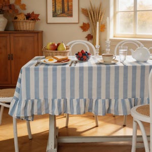 May include: A rectangular tablecloth featuring vertical light blue and white stripes with a ruffled hem. The fabric covers a dining table set with a plate of breakfast food, a bowl of mixed berries, a teacup, a glass of water, and a white teapot.