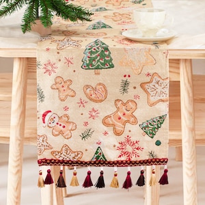 May include: Beige Christmas table runner with gingerbread men, Christmas trees, and snowflake patterns. The runner has a red border with tassels. A cup and saucer are on the table. The runner is on a wooden table.
