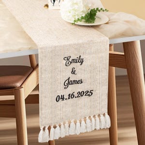 May include: Beige table runner with embroidered text reading "Emily & James" and the date "04.16.2025". The runner has a textured weave and white tassel fringe. A white floral arrangement sits on a white plate.