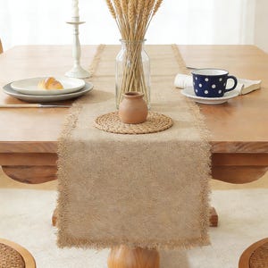 Puede incluir: Una mesa de comedor rústica con un camino de mesa de yute natural con bordes deshilachados. La mesa está decorada con un plato blanco, una taza azul con lunares, una pequeña olla de terracota y un jarrón con espigas de trigo, creando un ambiente cálido.