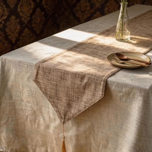 May include: A neutral-toned table setting featuring a beige tablecloth and a brown woven table runner. A small plate with wooden utensils sits on the runner. A glass vase with flowers and a tassel add decorative elements.
