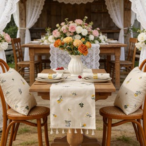 Può includere: Una scena di pranzo all'aperto splendidamente allestita con un tavolo di legno, sedie e composizioni floreali. Il tavolo è adornato con un runner floreale, piatti, tovaglioli e cuscini decorativi. Un vaso di rose arancioni e gialle è al centro.