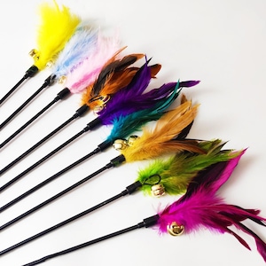May include: A collection of colorful cat toys with black handles and vibrant feathers. Each toy features a different color of feathers, including yellow, blue, pink, orange, purple, teal, green, and magenta. Small gold bells are attached to the base of the feathers.