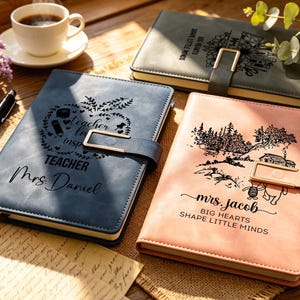Può includere: Tre quaderni personalizzati con chiusure dorate sono esposti su una superficie di legno. Uno è blu con "TEACHER Mrs. Daniel" e un design a mela. Un altro è rosa con "Mrs. Jacob" e una scena di foresta. Il terzo è grigio.