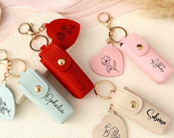 May include: A collection of personalized keychains in various colors, including red, light blue, pink, and beige. Each keychain features a rectangular case with a snap closure and a heart-shaped charm with a floral design. Names are printed on the cases.