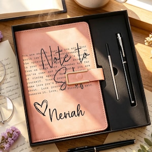 Könnte beinhalten: Ein pfirsichfarbenes Notizbuch mit goldfarbenem Verschluss, das den Text 'Note to self' und 'Neriah' sowie ein Herzsymbol zeigt. Das Notizbuch liegt in einer schwarzen Geschenkbox neben einem schwarzen Stift und einer Mine, mit einem Stift darunter.