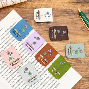 Op de afbeelding: Een verzameling kleurrijke leren bladwijzers met bloemmotieven en namen als "Heather", "Madison", "Deborah" en "Christina". Elke bladwijzer heeft een illustratie van een open boek. De bladwijzers liggen op een houten oppervlak met een pen en een boek.