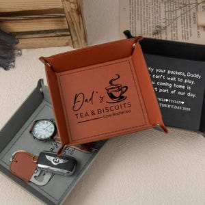 May include: A square tan leather valet tray with metal corner snaps, featuring a black printed graphic of a teacup and the text "Dad's TEA & BISCUITS Love Rachel xxx". A second black tray with white text and a grey tray with a watch and key fob.