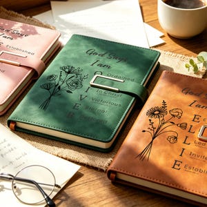 May include: Three leather journals in pink, green, and brown, each with a floral design and the words "God Says I Am" and other inspirational words. The journals have a strap closure and are on a wooden surface.