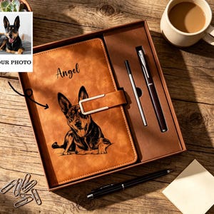 May include: A tan leather-effect journal featuring a black ink illustration of a dog and the name Angel printed in script. The journal has a magnetic strap closure and is presented in a gift box with a matching pen and a spare ink refill cartridge.