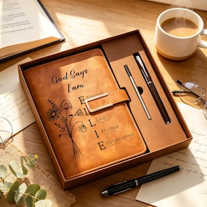 May include: A brown leather journal with the text "God Says I am" and "Eternel, Led, Invited, Established" is displayed in a gift box with two pens. A cup of coffee and a pair of glasses are also in the image.