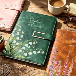Può includere: Un diario verde scuro con una chiusura in metallo dorato, caratterizzato da una stampa floreale di mughetti bianchi e dal testo "Sophia NEW CHAPTER" stampato sulla copertina, insieme ad altri diari, una tazza di caffè e occhiali.