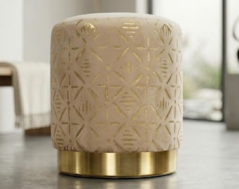 Elegancki, okrągły, beżowy, aksamitny puf z geometrycznym nadrukiem w kolorze złotej folii i metalową podstawą.