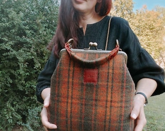 May include: A structured handbag featuring a brown and orange plaid wool pattern with a bronze-toned metal kiss-lock frame closure. The bag includes a small rectangular leather patch embossed with the letters AIM and a short handle.