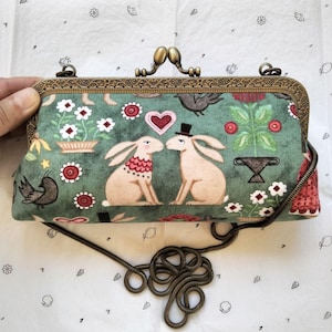 Puede incluir: Un bolso de mano rectangular con exterior de tela verde que presenta un estampado de conejos, pájaros, flores y corazones. El bolso tiene un marco de metal de bronce antiguo con cierre de boquilla y una cadena de metal a juego.