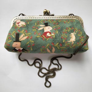 Puede incluir: Un bolso de mano rectangular con tela verde y un patrón repetitivo de conejos con sombreros de copa y chalecos rojos, rodeados de coronas florales. El bolso tiene un marco de metal en tono latón antiguo con cierre de clip y una cadena.