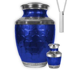 May include: A cobalt blue urn with a textured surface and silver accents. A smaller matching urn and a silver pendant necklace are also shown. The urns have a decorative pattern around the top and bottom.