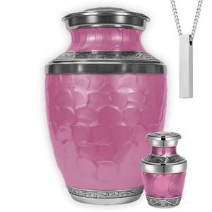 May include: A pink urn with a textured surface and silver accents, accompanied by a smaller matching urn and a silver pendant necklace. The urns have decorative patterns around the top and bottom.
