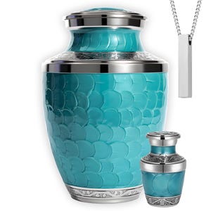 May include: A teal cremation urn with a textured, scale-like pattern, silver accents, and a matching miniature urn. A silver pendant necklace with a rectangular bar is also shown. The urns are designed for memorial purposes.
