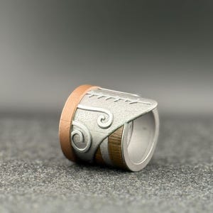 Puede incluir: Un anillo plateado con detalles en cobre y madera. El anillo presenta un dise&ntilde;o decorativo en la parte plateada, con una forma curva y un acabado liso y pulido. El anillo se muestra sobre una superficie gris.