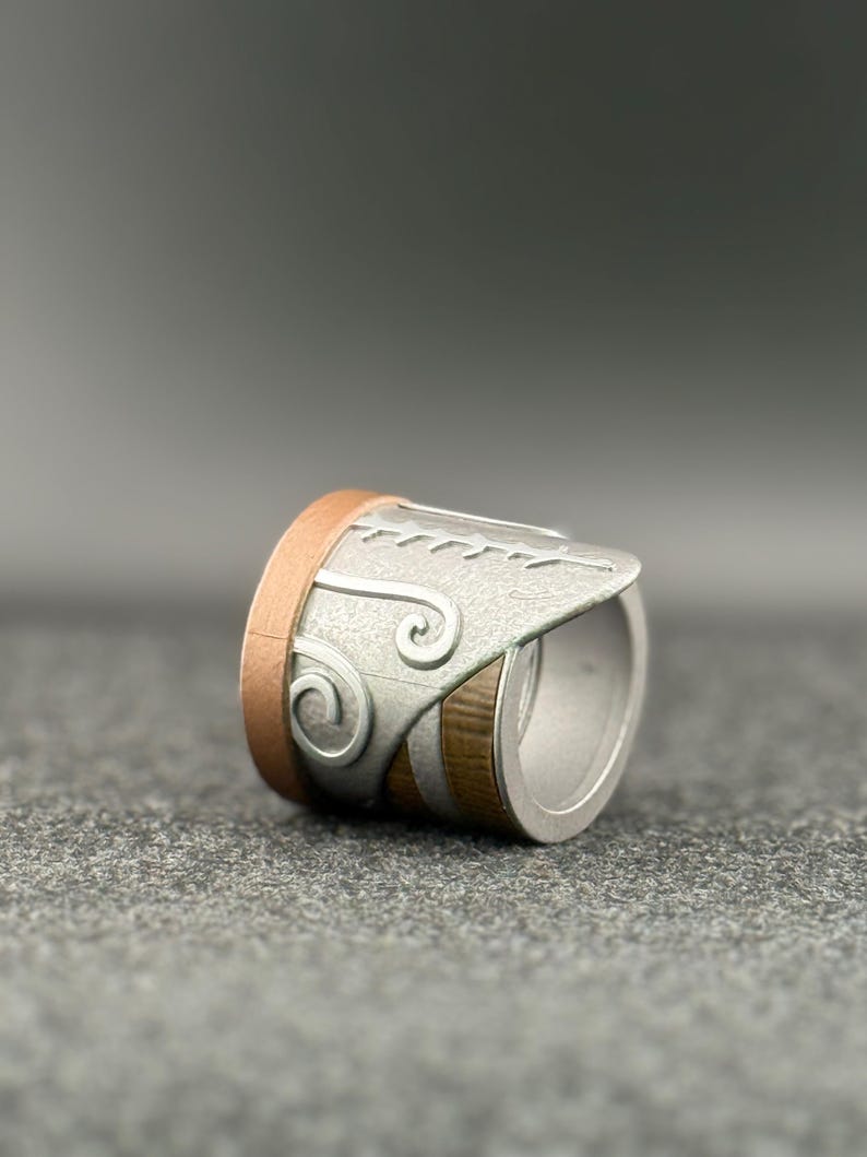 Puede incluir: Un anillo plateado con detalles en cobre y madera. El anillo presenta un dise&ntilde;o decorativo en la parte plateada, con una forma curva y un acabado liso y pulido. El anillo se muestra sobre una superficie gris.