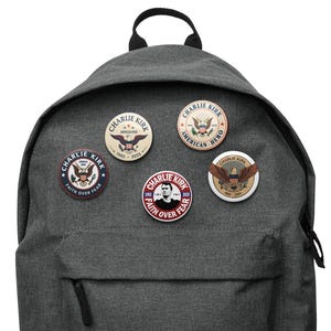 May include: A gray backpack with five round pinback buttons attached. The buttons feature the text "Charlie Kirk" and various patriotic designs, including eagles and the words "American Hero" and "Faith Over Fear."