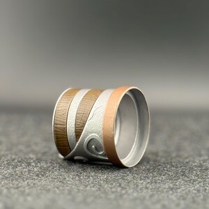 Puede incluir: Un anillo cil&iacute;ndrico con bandas alternas de veta de madera marr&oacute;n, plata y metal color cobre. La banda plateada presenta un dise&ntilde;o de remolino decorativo. El interior del anillo es de color plateado liso.