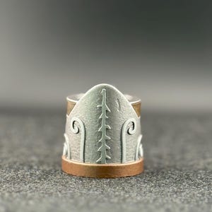 Puede incluir: Un anillo de plata con un dise&ntilde;o detallado. El anillo presenta una banda de plata texturizada con detalles de color cobre en la parte superior e inferior. El dise&ntilde;o incluye una l&iacute;nea vertical central y patrones curvos en los lados.