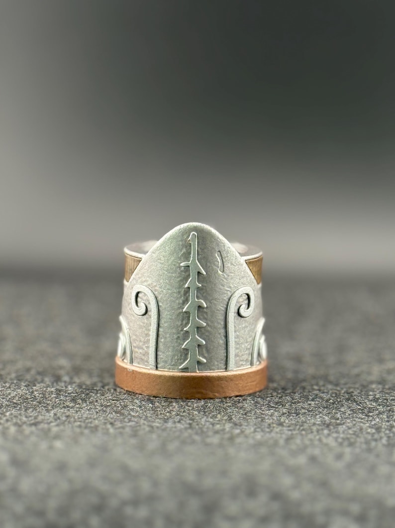 Puede incluir: Un anillo de plata con un dise&ntilde;o detallado. El anillo presenta una banda de plata texturizada con detalles de color cobre en la parte superior e inferior. El dise&ntilde;o incluye una l&iacute;nea vertical central y patrones curvos en los lados.