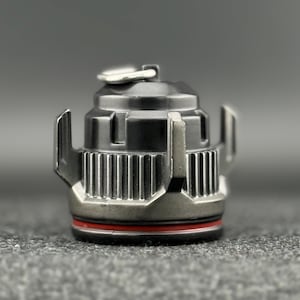 May include: A close-up of a metallic, industrial-style object. The object is primarily dark gray with silver accents and a red band. It has a cylindrical body with vertical grooves and protruding side elements. A small, silver loop is visible on top.