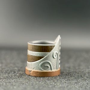 Puede incluir: Un anillo decorativo con un dise&ntilde;o plateado, marr&oacute;n y cobrizo. El anillo presenta una textura similar a la madera en la banda, detalles plateados con un dise&ntilde;o de remolino y una base de cobre. El anillo es una joya &uacute;nica.