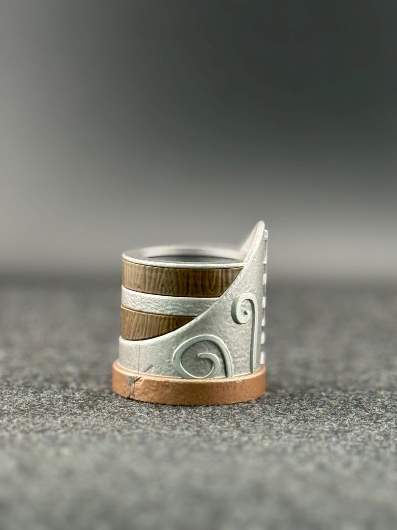 Puede incluir: Un anillo decorativo con un dise&ntilde;o plateado, marr&oacute;n y cobrizo. El anillo presenta una textura similar a la madera en la banda, detalles plateados con un dise&ntilde;o de remolino y una base de cobre. El anillo es una joya &uacute;nica.