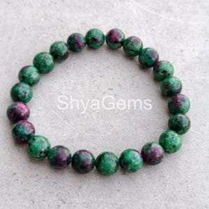 Puede incluir: Un brazalete de cuentas con piedras preciosas redondas en tonos verdes y granates. El brazalete está hecho de múltiples cuentas ensartadas, formando una forma circular. El texto "ShyaGems" es visible en la superficie.
