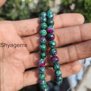 May include: Two strands of gemstone beads. The beads are a mix of green and magenta colors. The beads are round and appear to be polished. The text "Shyagems" is in the lower left corner.