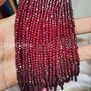 Puede incluir: Primer plano de hebras de cuentas facetadas de color rojo intenso. Las cuentas están ensartadas, creando un efecto en cascada. Las cuentas son de un color rojo oscuro intenso, y las facetas captan la luz, creando un efecto brillante. El texto "Shya Gems" es visible.