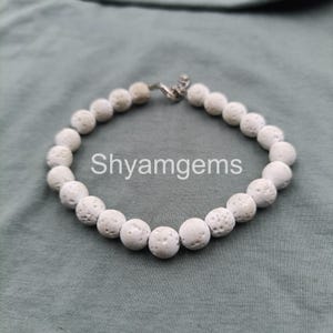 Puede incluir: Una pulsera de cuentas blancas con un cierre plateado. La pulsera está hecha de cuentas redondas y porosas. El texto "Shyamgems" está impreso en el fondo.