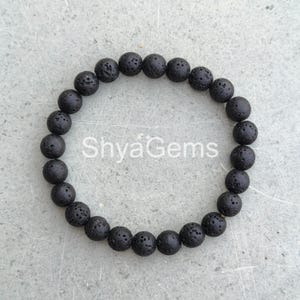 Puede incluir: Una pulsera de cuentas de piedra de lava negra. La pulsera está hecha de cuentas redondas y porosas ensartadas. Las cuentas son de color negro mate oscuro. El texto "ShyaGems" es visible en la imagen.