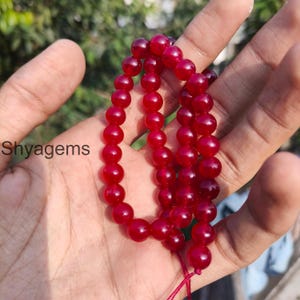 May include: A strand of deep red, polished gemstone beads. The beads are round and strung together, with a red string visible at the bottom. The text "Shyagems" is visible in the lower left corner.