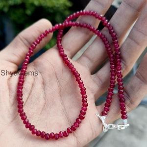 May include: A necklace of small, faceted ruby red beads, secured with a silver clasp. The beads are closely strung together, creating a continuous line of colour. The necklace is shown against a neutral background.