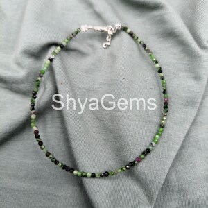 May include: A delicate beaded necklace featuring small, round gemstones in shades of green, black, and red. The necklace is strung on a fine chain and has a silver clasp. The text "ShyaGems" is visible in the image.