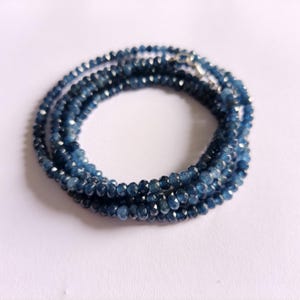 Puede incluir: Un collar compuesto por múltiples hilos de pequeñas piedras preciosas facetadas de color azul oscuro. Los hilos están dispuestos en un patrón circular, con un cierre plateado visible en la parte superior. Las piedras preciosas tienen un aspecto brillante.